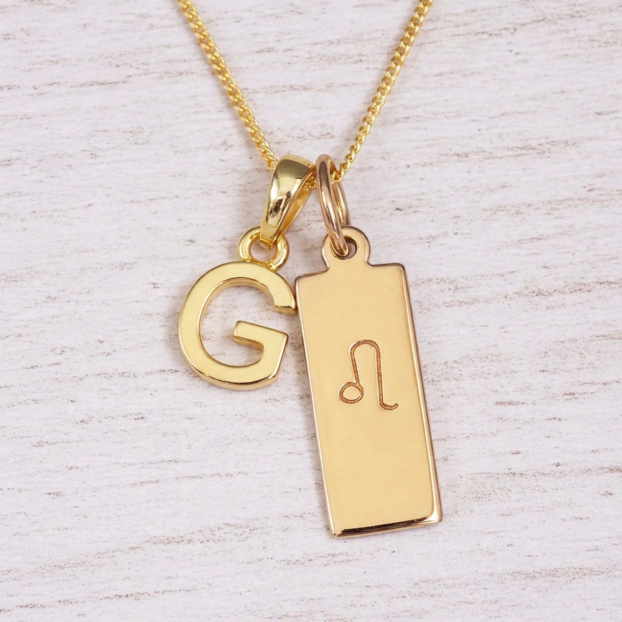 Gold initial and Leo zodiac necklace with a letter G pendant and engraved Leo bar on white wood, close-up