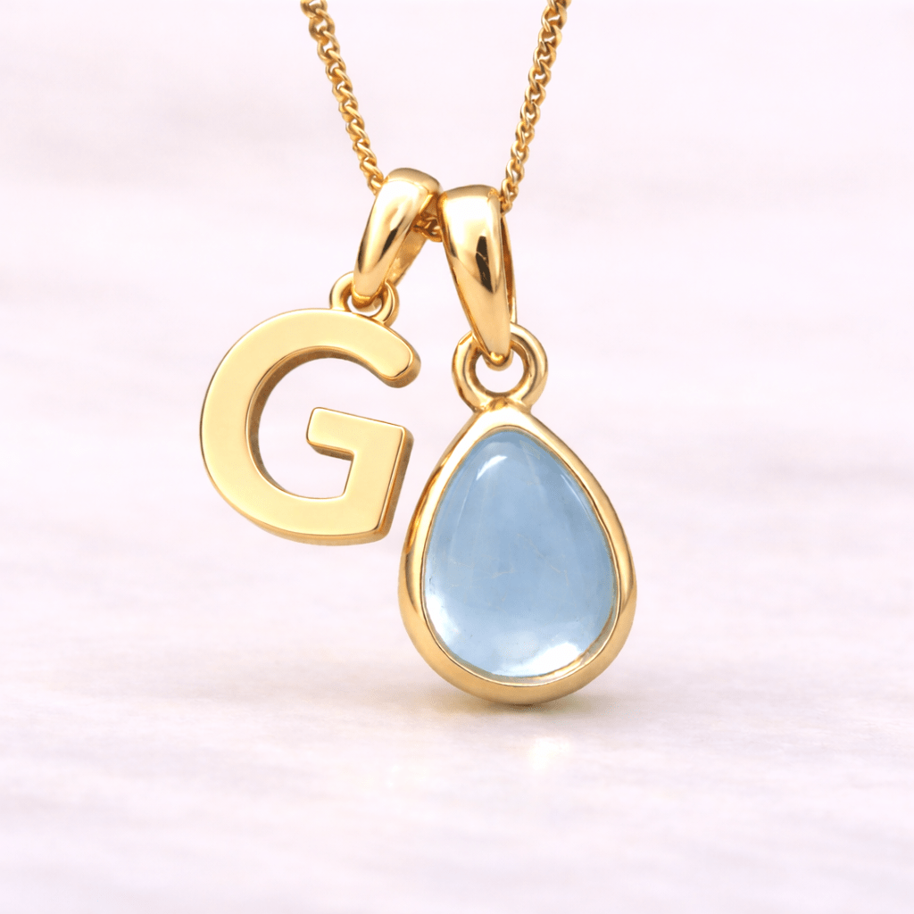 Gold necklace with 'G' pendant and teardrop-shaped blue topaz gemstone on a light pink background