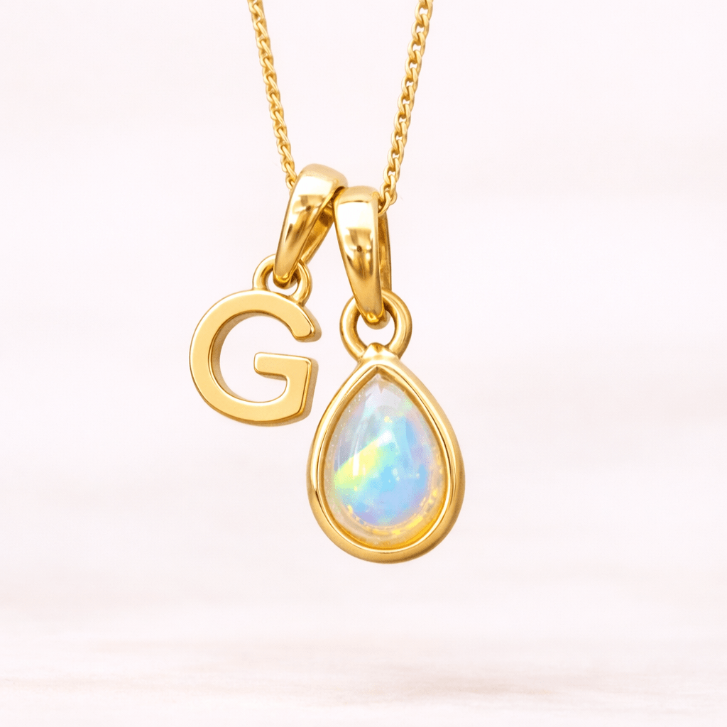 Gold necklace with 'G' pendant and teardrop-shaped opal gem on a light pink background