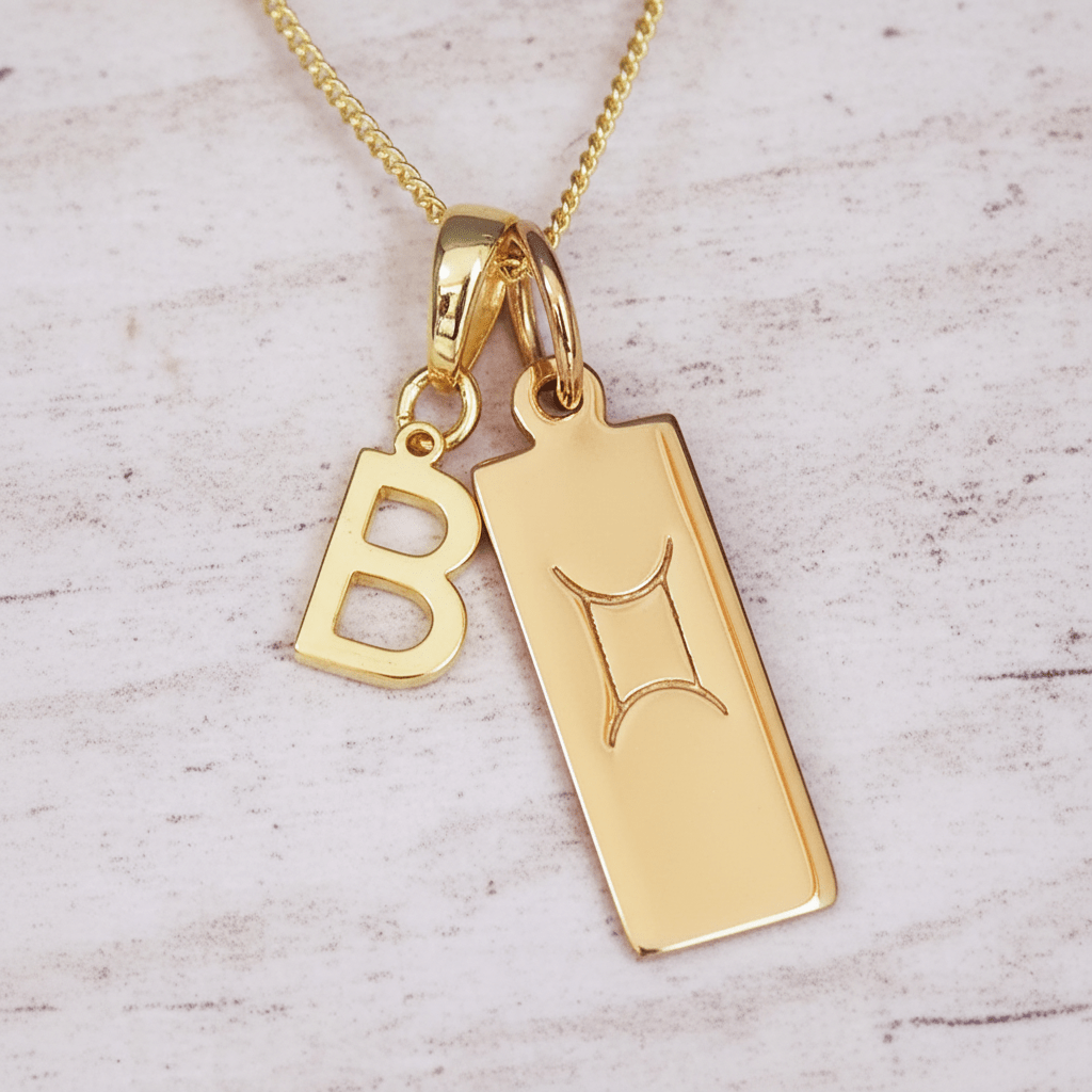 Gold necklace with a 'B' initial pendant and Gemini zodiac pendant symbol on a light wooden background.