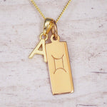 Gold necklace with a 'A' initial pendant and Gemini zodiac pendant symbol on a light wooden background.
