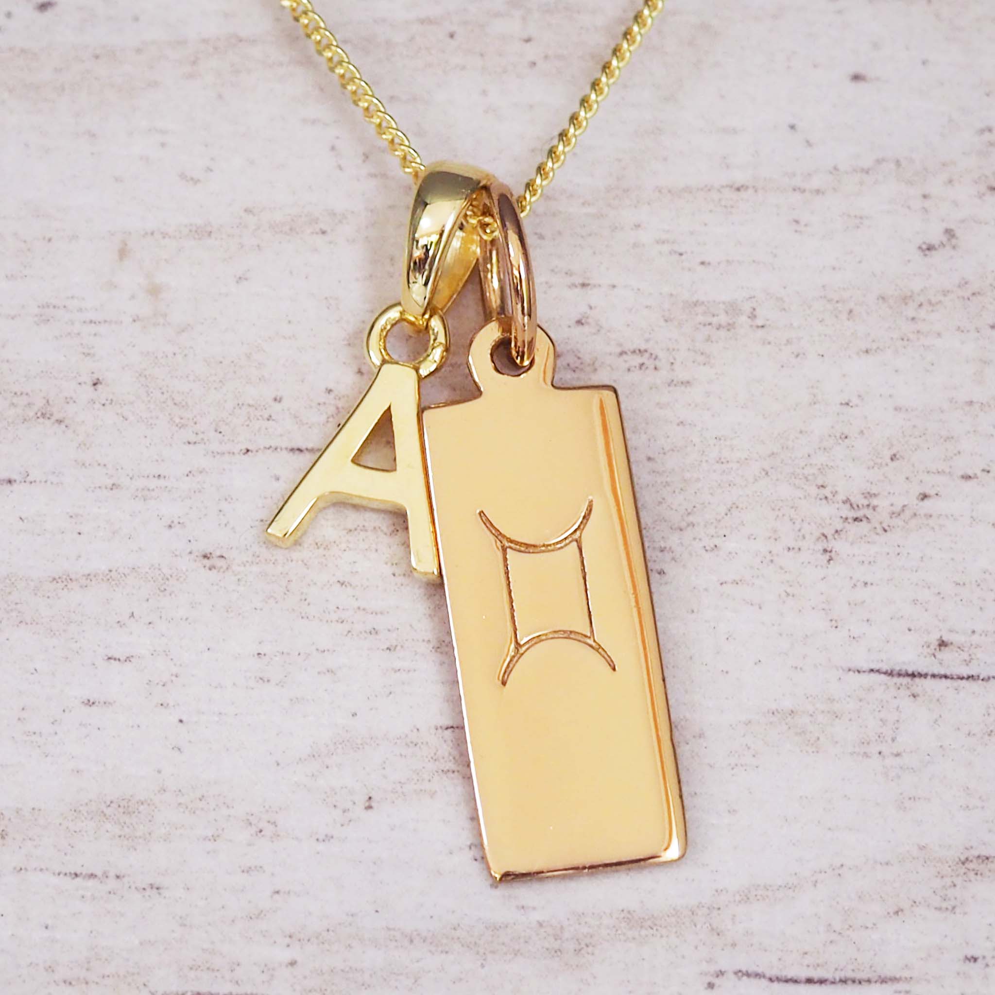 Gold necklace with a 'A' initial pendant and Gemini zodiac pendant symbol on a light wooden background.