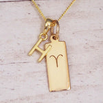 Gold initial H and Aries zodiac bar necklace, close-up of the letter charm and engraved pendant on whitewashed wood