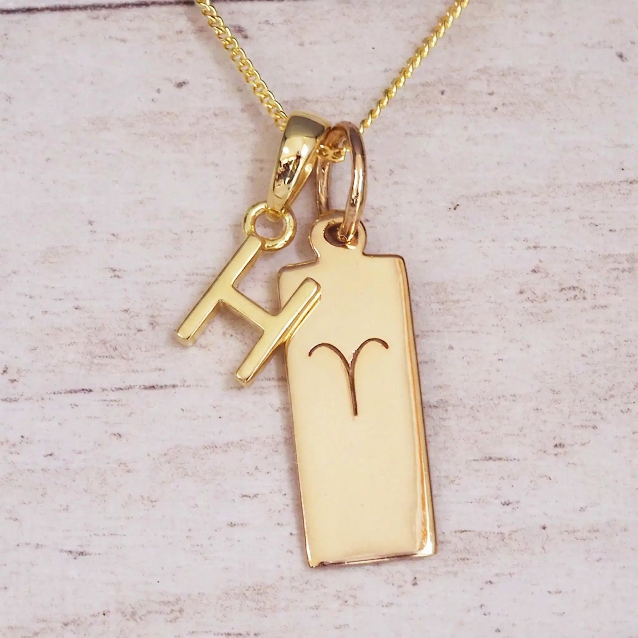 Gold initial H and Aries zodiac bar necklace, close-up of the letter charm and engraved pendant on whitewashed wood