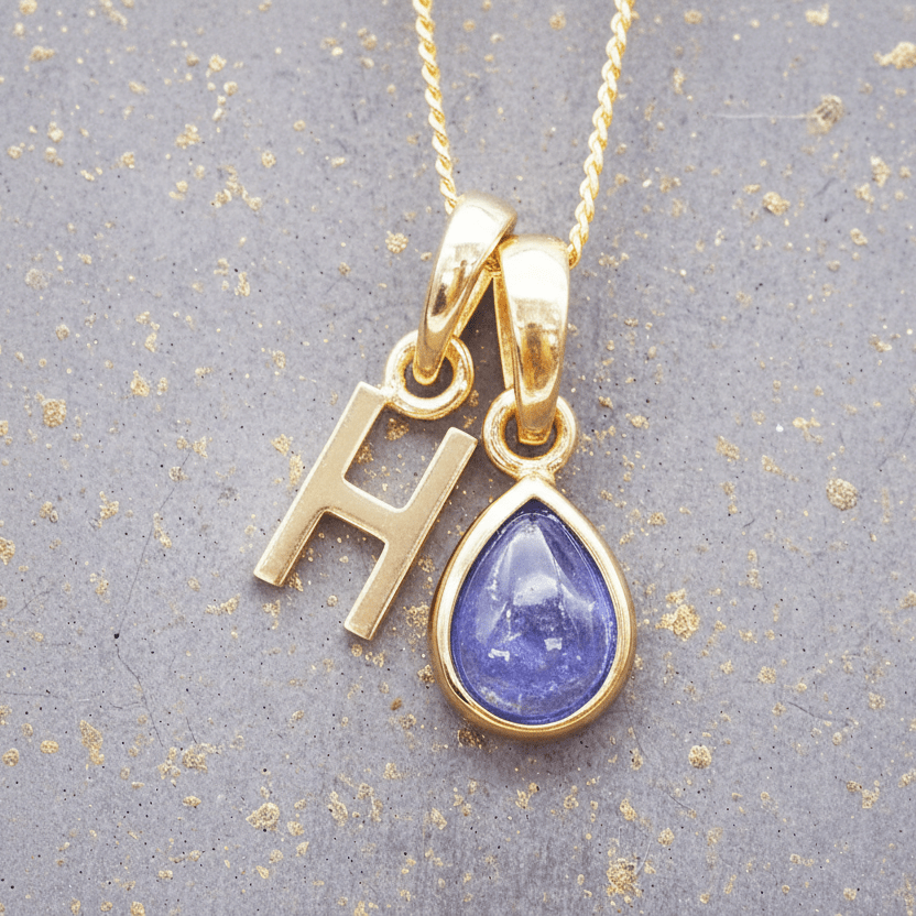 Gold necklace with 'H' pendant and teardrop-shaped purple tanzanite gemstone on a textured gray background