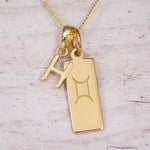Gold initial H and Gemini zodiac bar necklace, close-up of the letter charm and engraved pendant on whitewashed wood