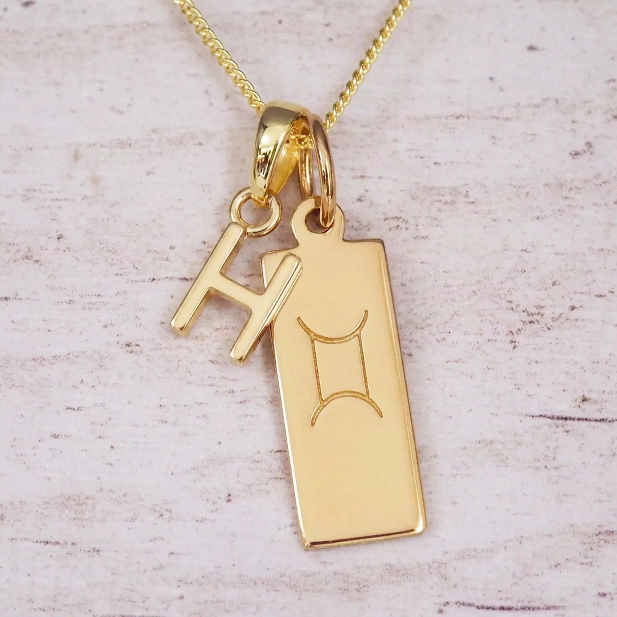 Gold initial H and Gemini zodiac bar necklace, close-up of the letter charm and engraved pendant on whitewashed wood