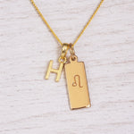 Gold initial and Leo zodiac necklace with a letter H pendant and engraved Leo bar on white wood, close-up