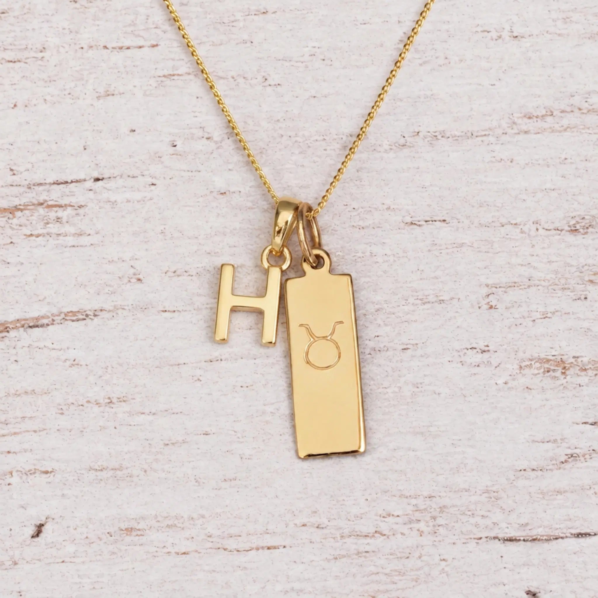 Gold H Initial and Taurus Necklace on a fine gold chain against a light wooden background