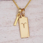 Gold initial I and Aries zodiac bar necklace, close-up of the letter charm and engraved pendant on whitewashed wood