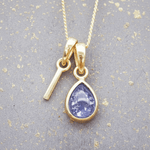 Gold necklace with a 'I' initial and a teardrop-shaped purple december birthstone natural tanzanite gemstone pendant on a textured gray background.