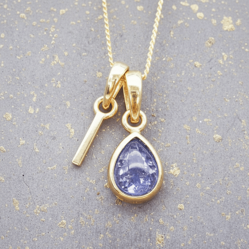 Gold necklace with a 'I' initial and a teardrop-shaped purple december birthstone natural tanzanite gemstone pendant on a textured gray background.