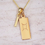 Gold initial I and Gemini zodiac bar necklace, close-up of the letter charm and engraved pendant on whitewashed wood