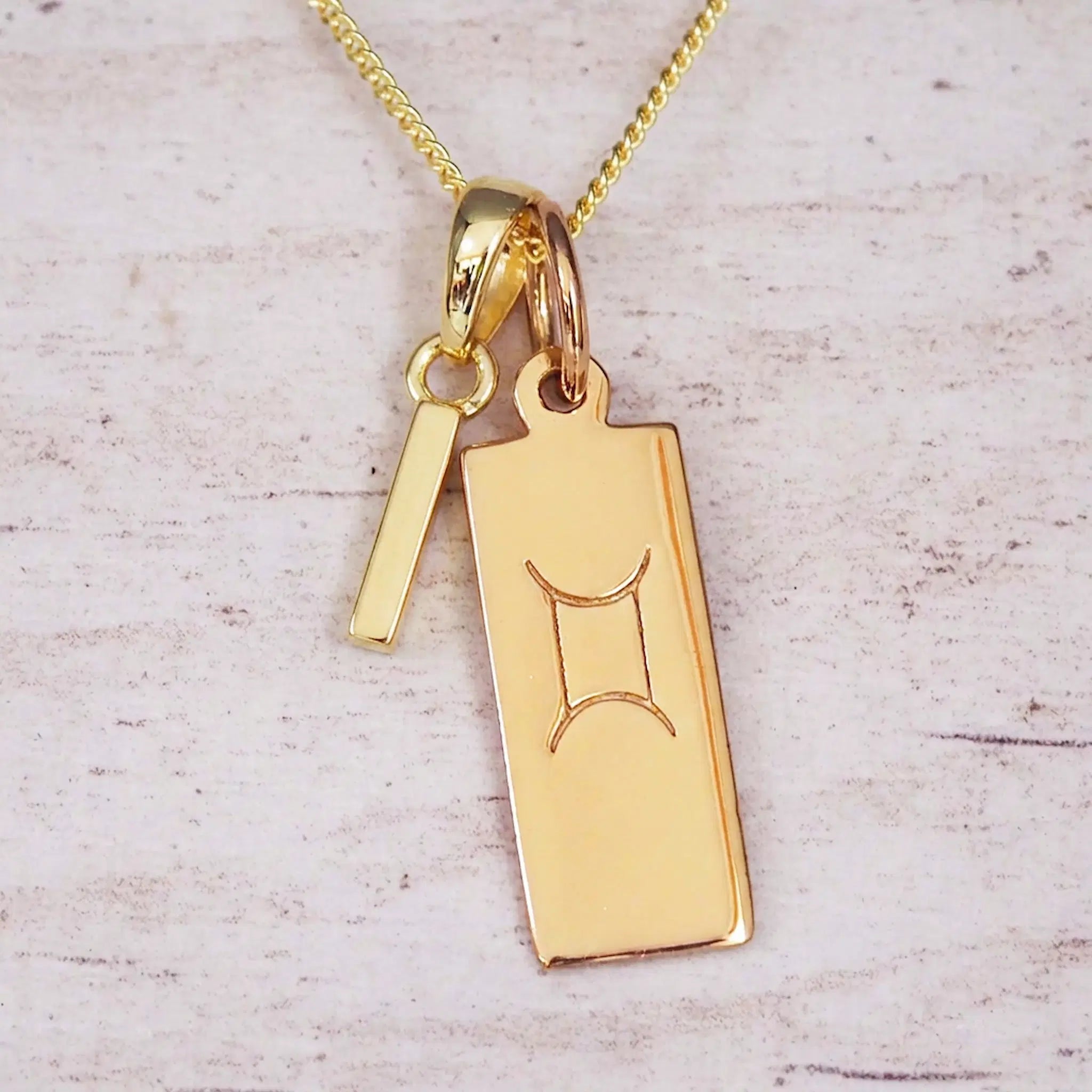 Gold initial I and Gemini zodiac bar necklace, close-up of the letter charm and engraved pendant on whitewashed wood