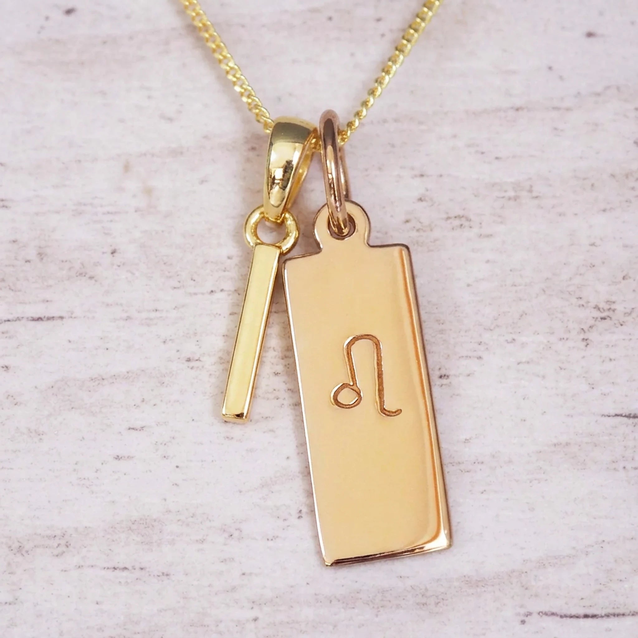 Gold initial and Leo zodiac necklace with a letter I pendant and engraved Leo bar on white wood, close-up