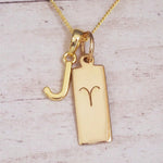 Gold initial J and Aries zodiac bar necklace, close-up of the letter charm and engraved pendant on whitewashed wood