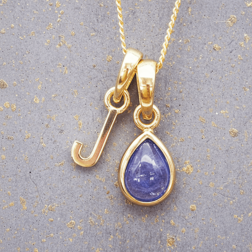 Gold J initial pendant with a purple tanzanite gemstone on a textured gray background