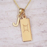 Gold initial J and Gemini zodiac bar necklace, close-up of the letter charm and engraved pendant on whitewashed wood