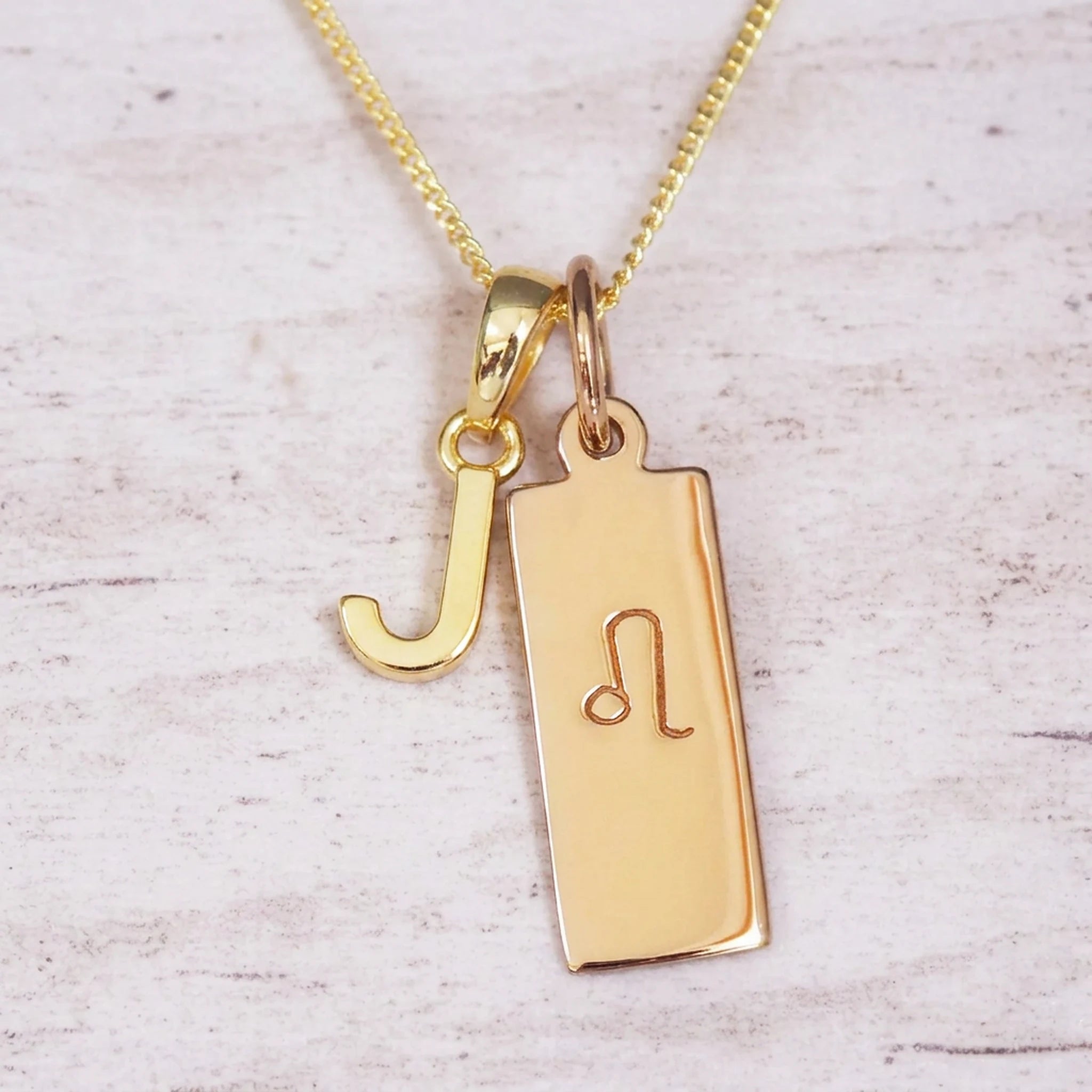 Gold initial and Leo zodiac necklace with a letter J pendant and engraved Leo bar on white wood, close-up