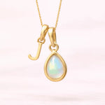Gold October birthstone necklace with a pear-shaped natural opal pendant and letter 'J' charm with a light background.
