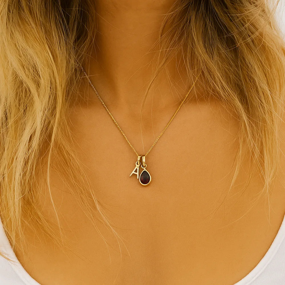 Gold initial A necklace with a teardrop-shaped garnet pendant charm worn by a person with blonde hair.
