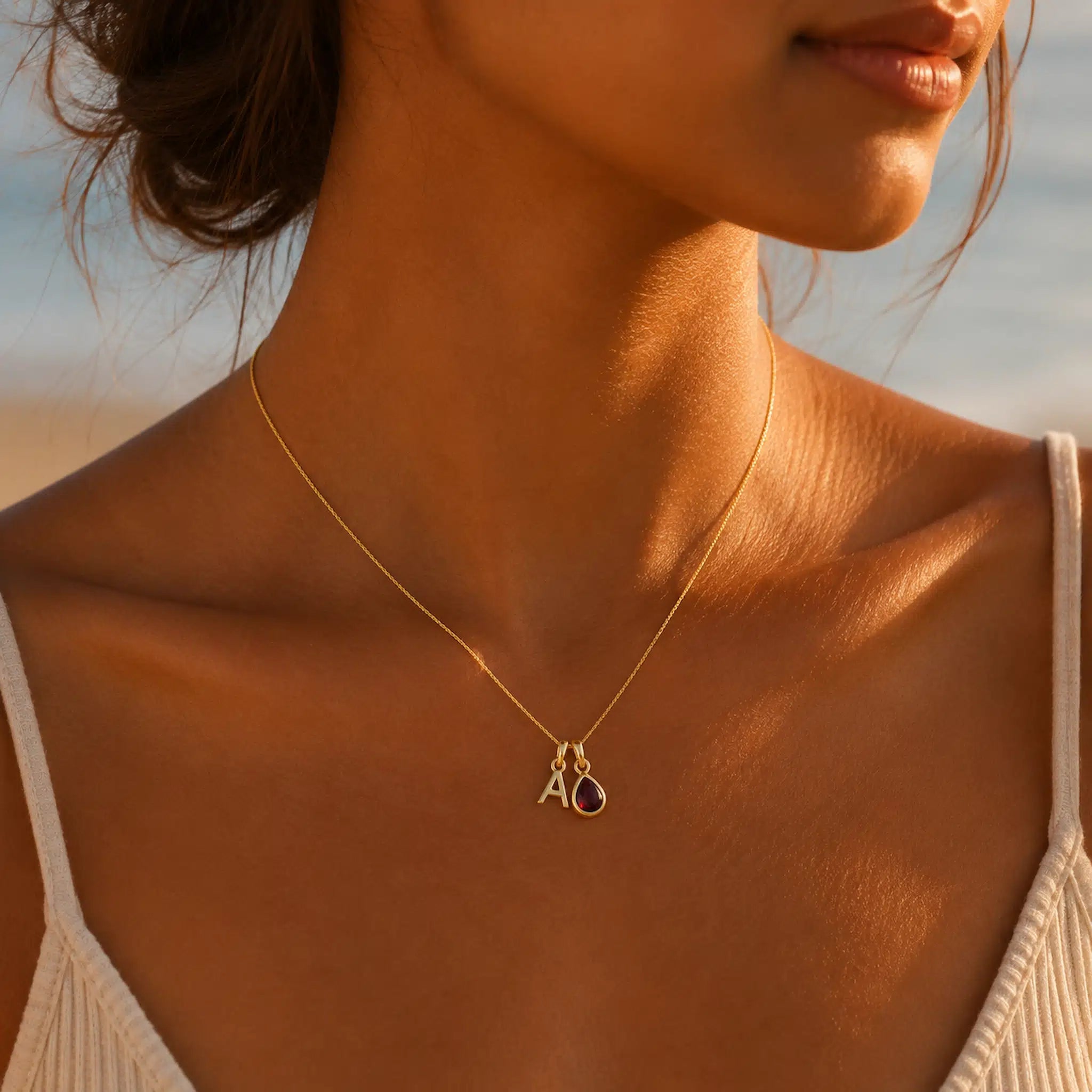 Woman wearing a gold necklace with a pendant featuring initials and a heart on a blurred beach background