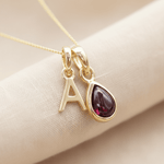 Gold necklace with a teardrop-shaped gemstone and letter 'A' pendant on a beige fabric background