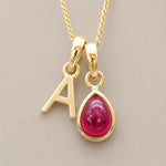 Gold July birthstone necklace with a pear-shaped natural ruby gemstone pendant and letter 'A' charm on a light background.