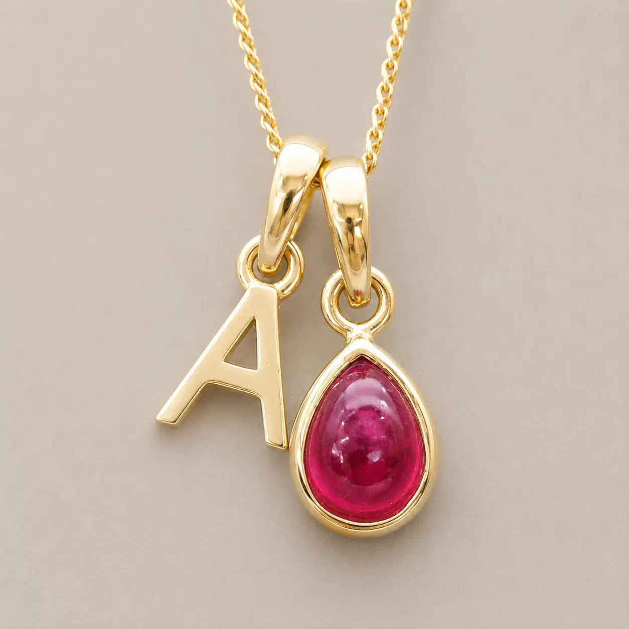 Gold July birthstone necklace with a pear-shaped natural ruby gemstone pendant and letter 'A' charm on a light background.