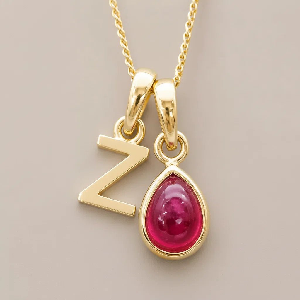 Gold necklace with 'Z' pendant and red natural ruby teardrop-shaped gemstone on a beige background