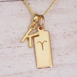 Gold initial K and Aries zodiac bar necklace, close-up of the letter charm and engraved pendant on whitewashed wood