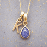 Gold necklace with 'K' initial pendant and tanzanite teardrop charm on a textured gray background