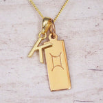 Gold initial K and Gemini zodiac bar necklace, close-up of the letter charm and engraved pendant on whitewashed wood
