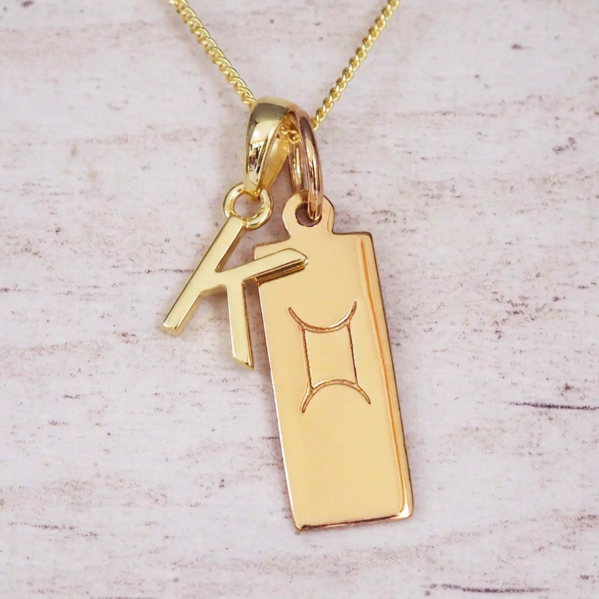 Gold initial K and Gemini zodiac bar necklace, close-up of the letter charm and engraved pendant on whitewashed wood