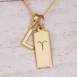 Gold initial L and Aries zodiac bar necklace, close-up of the letter charm and engraved pendant on whitewashed wood