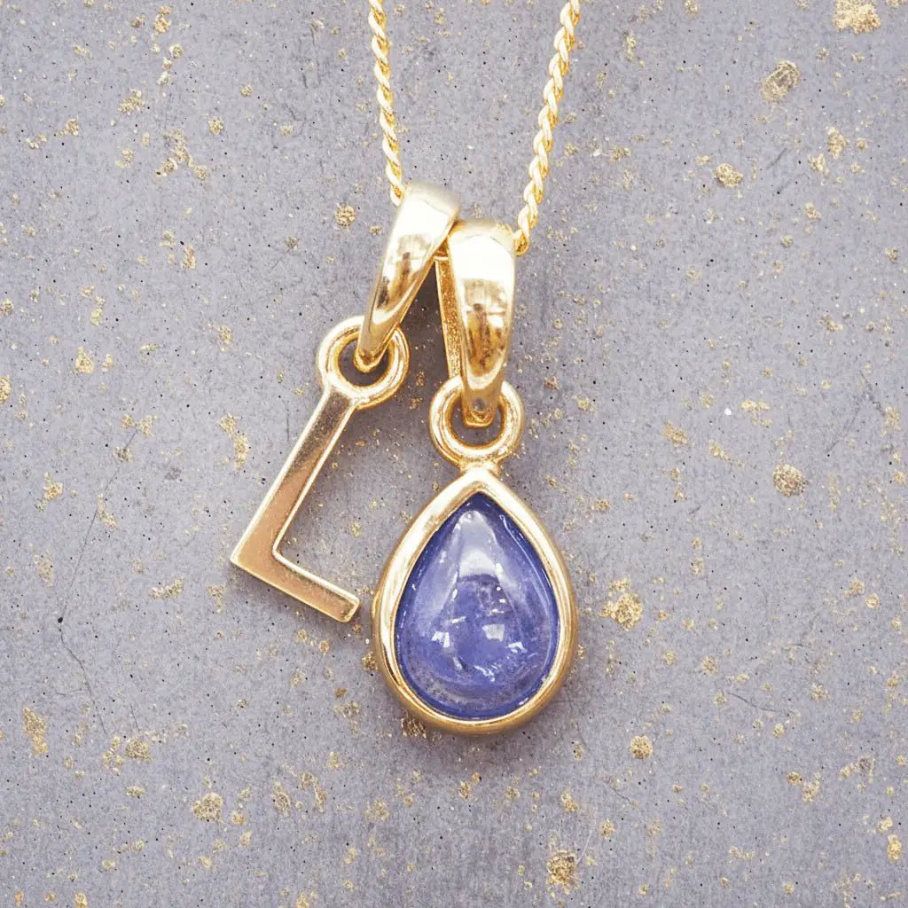 Gold December birthstone necklace with 'L' initial pendant and a natural tanzanite gemstone pendant on a grey background.