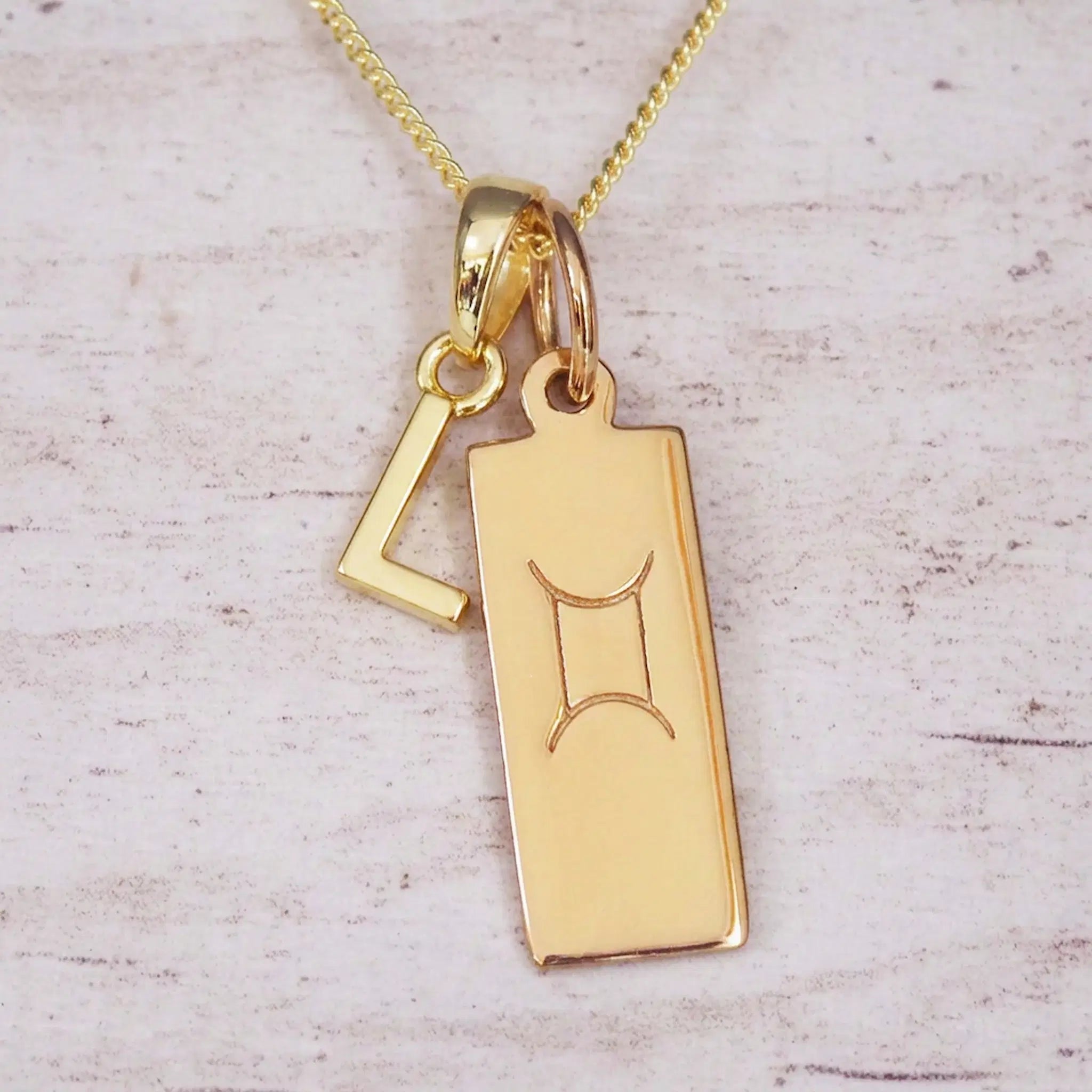 Gold initial L and Gemini zodiac bar necklace, close-up of the letter charm and engraved pendant on whitewashed wood