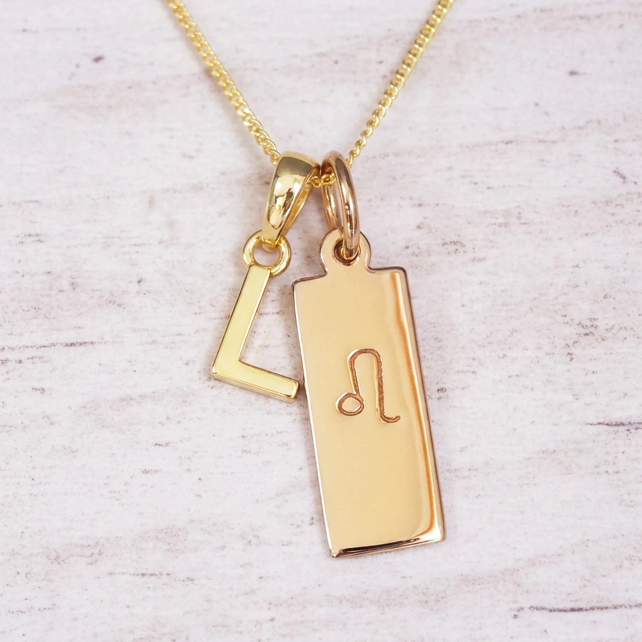 Gold initial and Leo zodiac necklace with a letter L pendant and engraved Leo bar on white wood, close-up