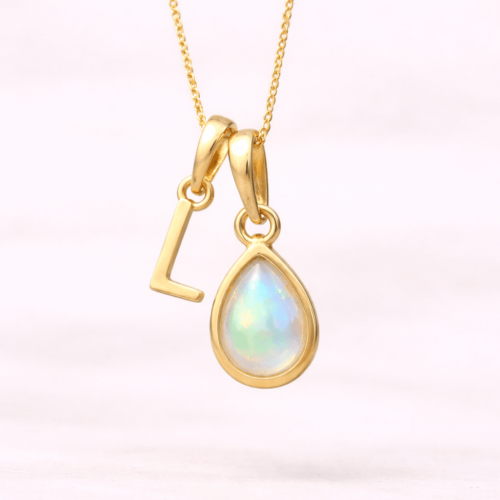 Gold necklace with a teardrop-shaped opal pendant and 'L' initial on a light pink background