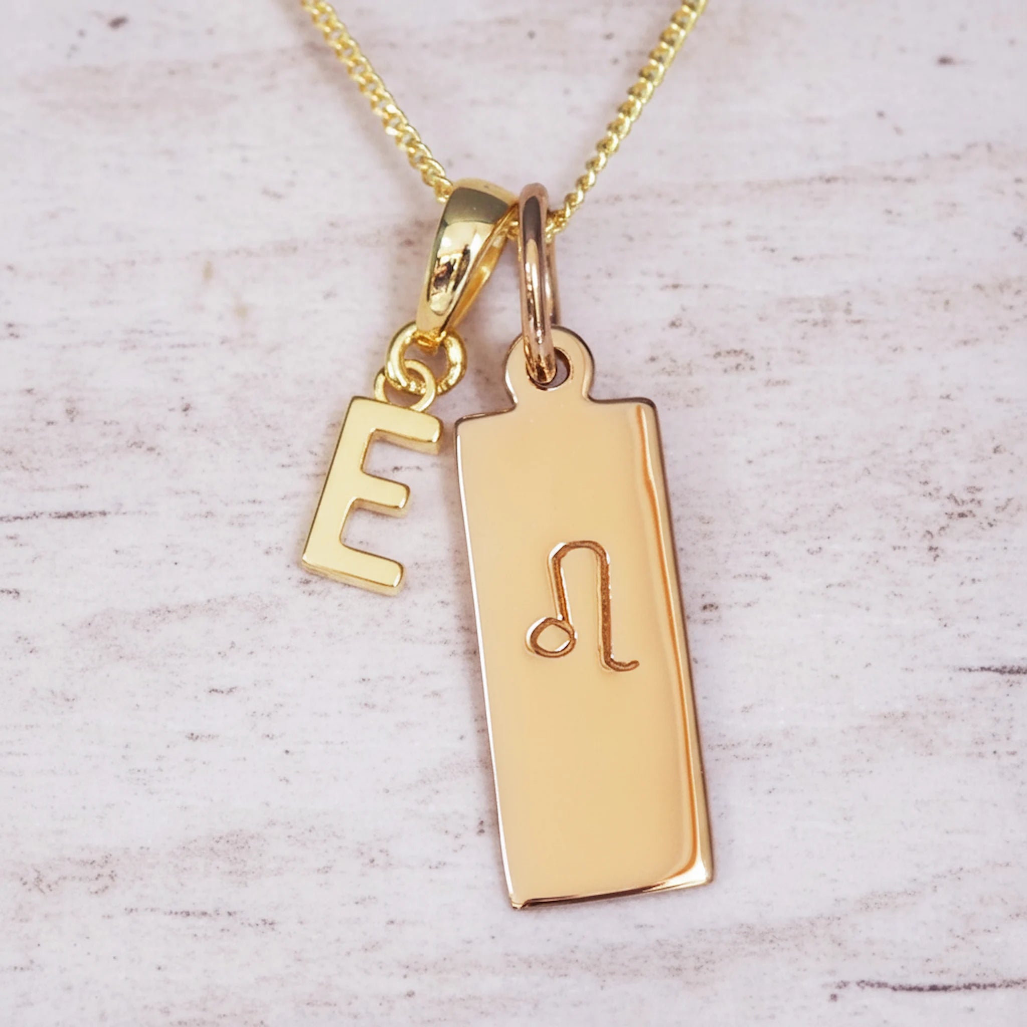 Close-up of personalised gold double-pendant necklace with E initial charm and polished rectangular tag engraved with the Leo zodiac glyph on a fine chain