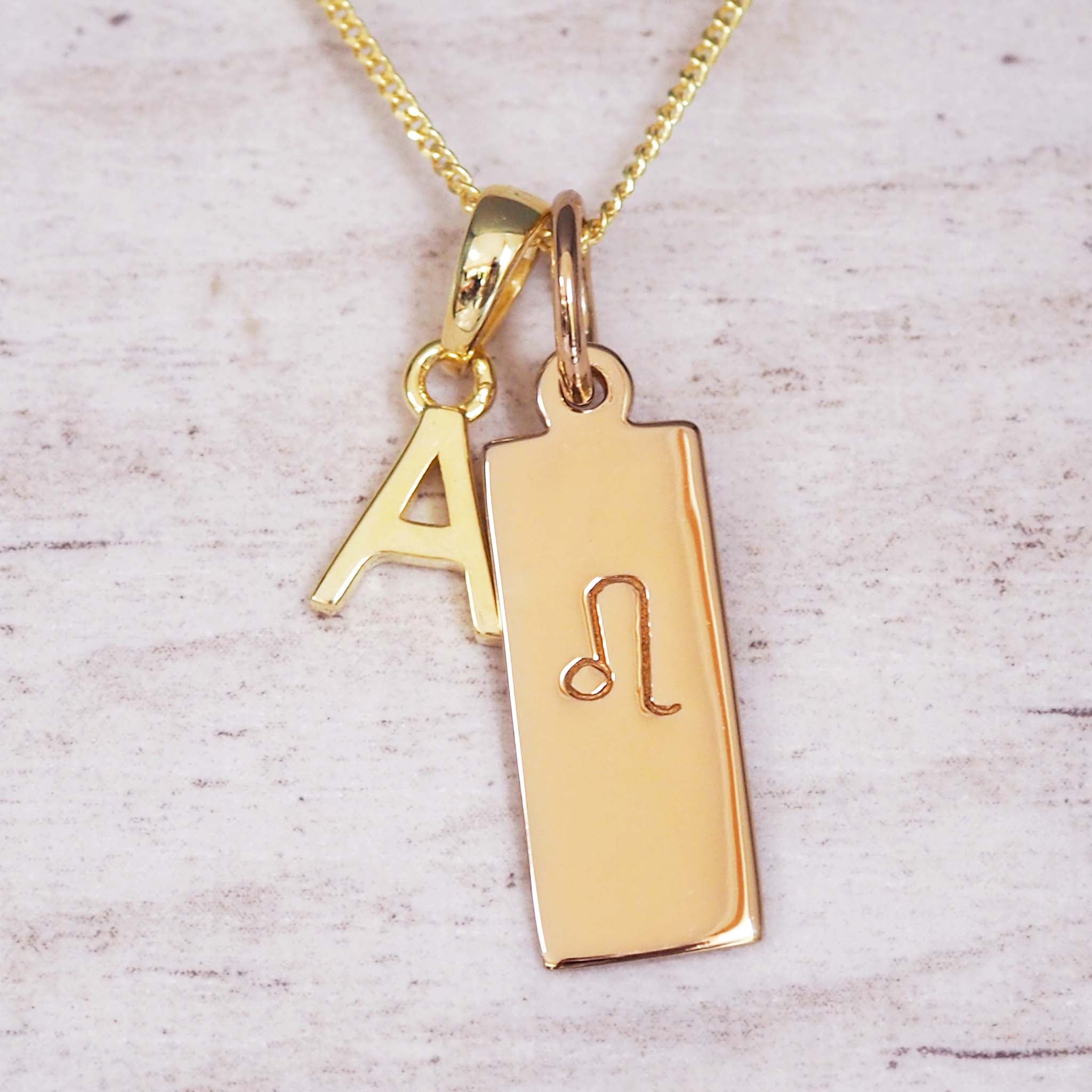 Close-up of personalised gold necklace with A initial charm and rectangular tag engraved with the Leo zodiac glyph