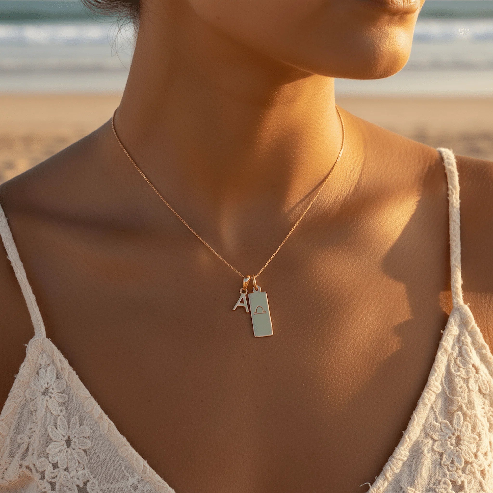 Woman on beach wearing a gold fine cable chain with two pendants. A initial 'A' pendant and a libra glyph rectangle pendant.
