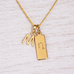 A gold initial necklace featuring a hanging letter 'M' and a rectangular tag pendant engraved with the capricorn zodiac symbol, displayed on a light wood background.