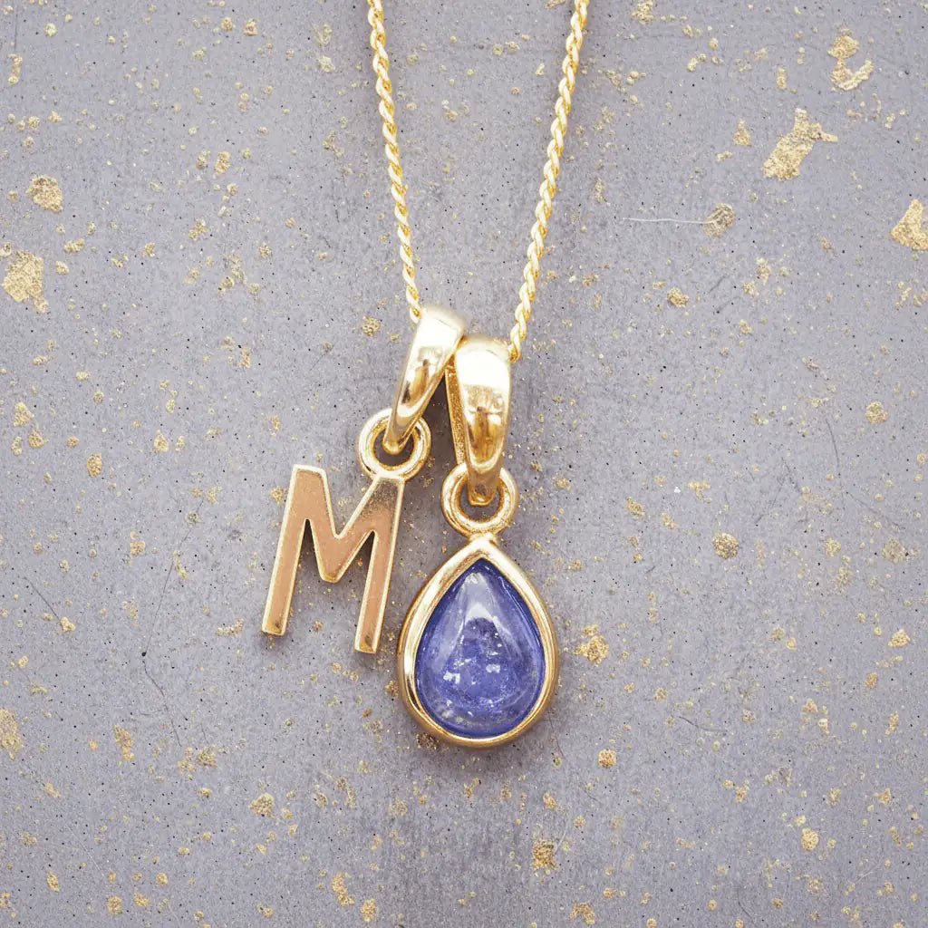 Gold December birthstone necklace with 'M' initial pendant and a natural tanzanite gemstone pendant on a grey background.