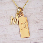 Gold initial M and Gemini zodiac bar necklace, close-up of the letter charm and engraved pendant on whitewashed wood
