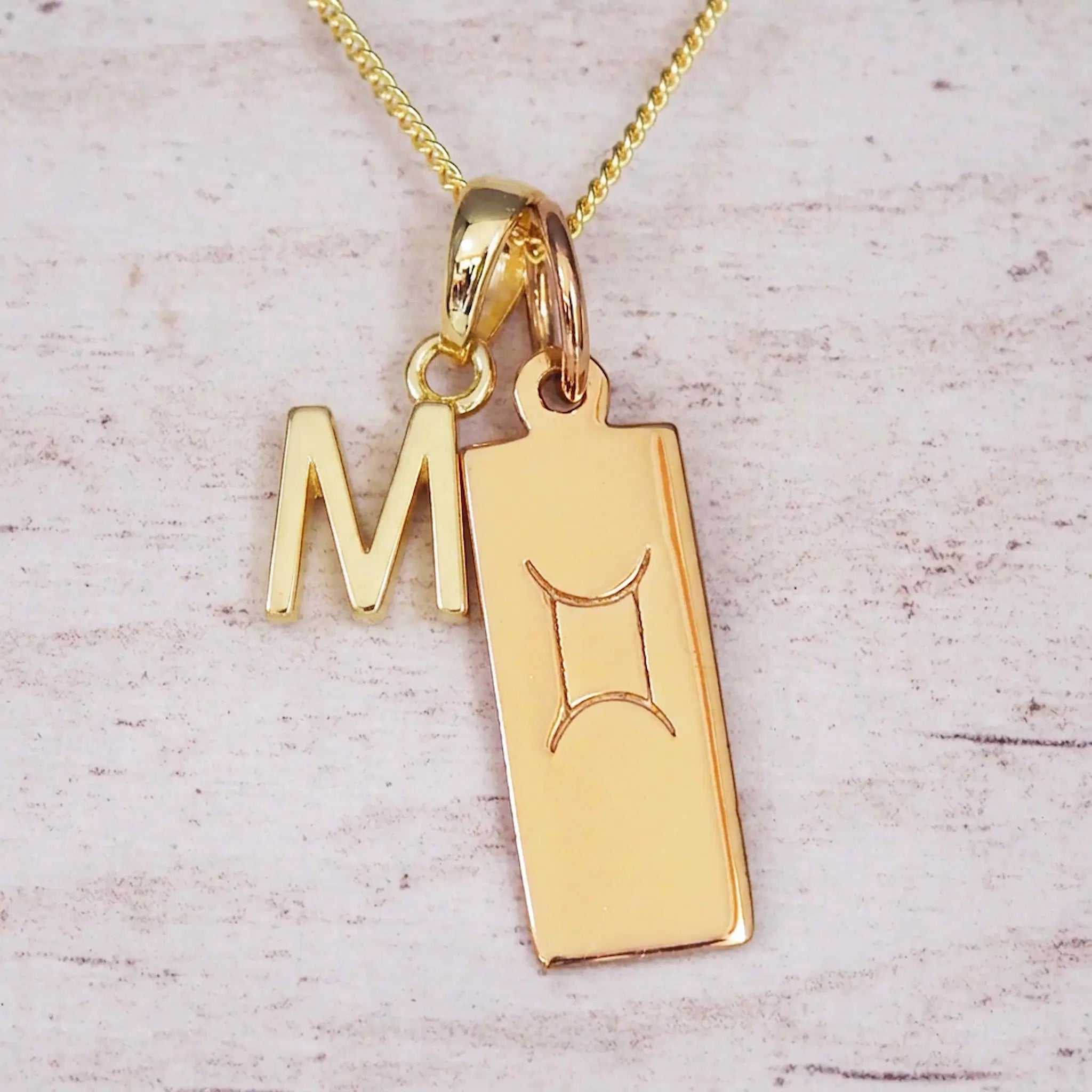 Gold initial M and Gemini zodiac bar necklace, close-up of the letter charm and engraved pendant on whitewashed wood