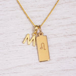 Gold initial and Leo zodiac necklace with a letter M pendant and engraved Leo bar on white wood, close-up