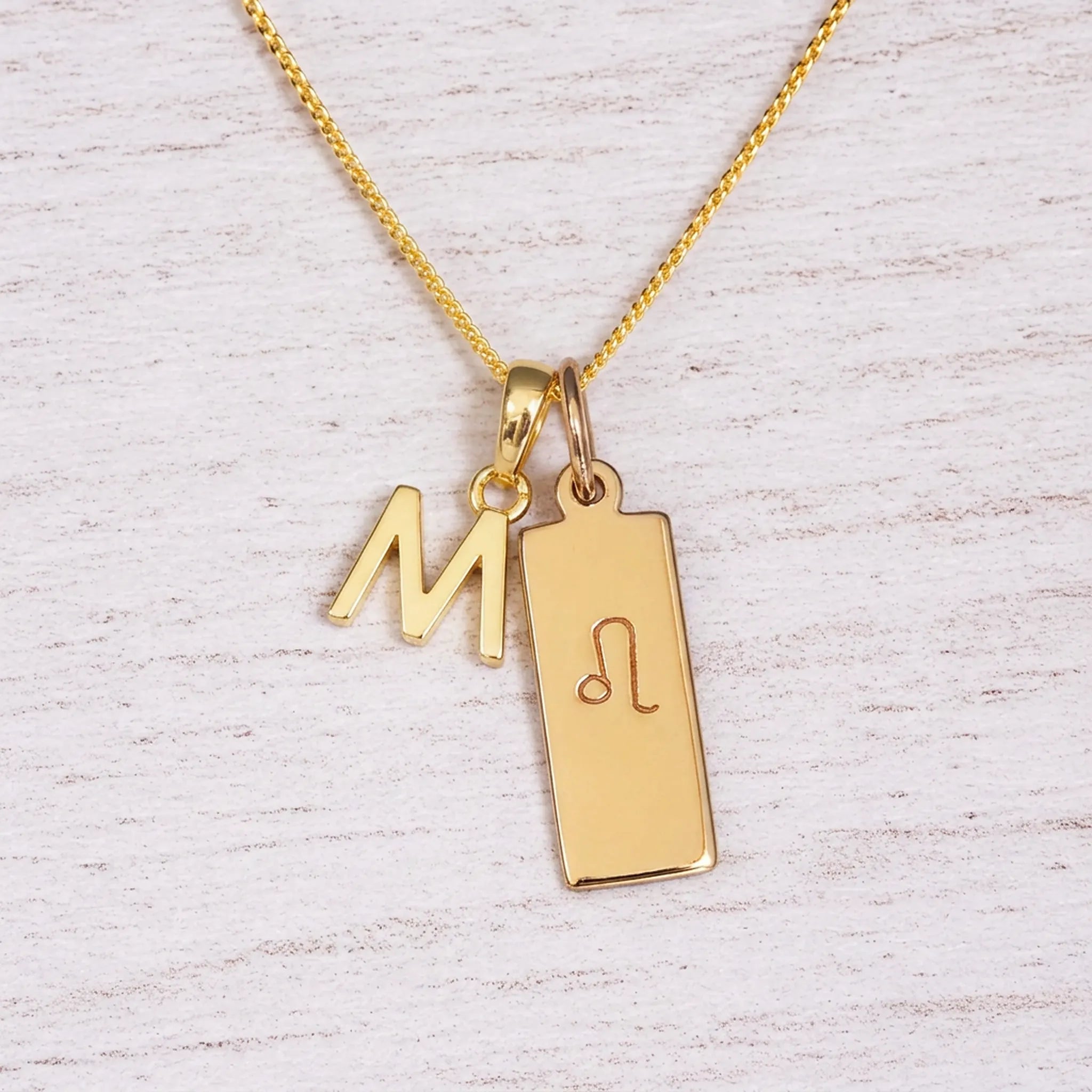 Gold initial and Leo zodiac necklace with a letter M pendant and engraved Leo bar on white wood, close-up