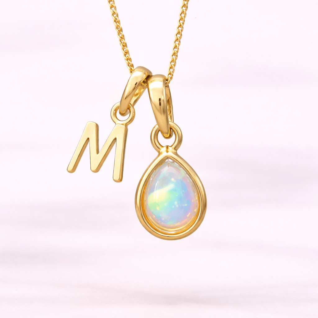 Gold necklace with 'M' pendant and teardrop-shaped opal stone on a light pink background
