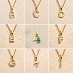 A 3x3 grid showing nine gold initial necklaces with letter pendants from A to I, displayed on a textured beige background. The center necklace features a gold letter A pendant paired with a green teardrop gemstone.